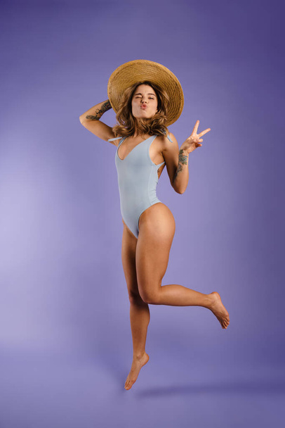 Full length of a happy young white woman wearing swimsuit and summer hat jumping over blue background - Photo, Image