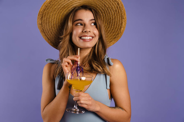 Young white woman in swimsuit and straw hat drinking cocktail isolated over purple background - Foto, Imagem