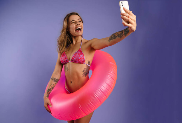 Young white woman in swimsuit taking selfie photo with rubber ring isolated over purple background - Photo, Image