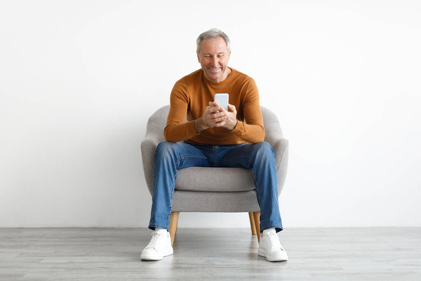 Портрет усміхненого зрілого чоловіка за допомогою смартфона, що сидить на стільці
 - Фото, зображення