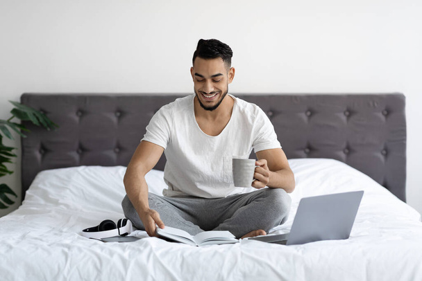 Arábiai férfi portréja, amint laptoppal, jegyzetfüzetekkel és kávéval ül az ágyban. - Fotó, kép