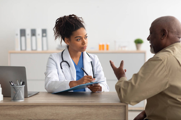 Νεαρός μαύρος γιατρός που εξηγεί το σχέδιο θεραπείας στον άνδρα ασθενή - Φωτογραφία, εικόνα