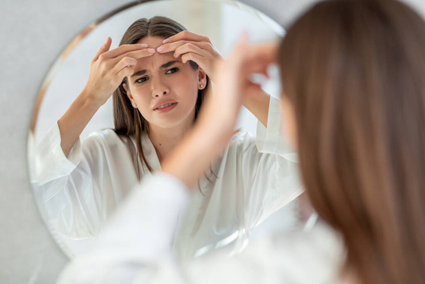Problema alla pelle. sconvolto giovane femmina poppante brufolo sulla fronte vicino specchio - Foto, immagini