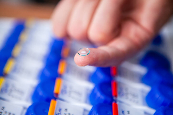 An optician doctor shows a new clear and clean contact lens resting on a finger. In the background are many packs of other new contact lens samples of different diopters and designs - Foto, afbeelding