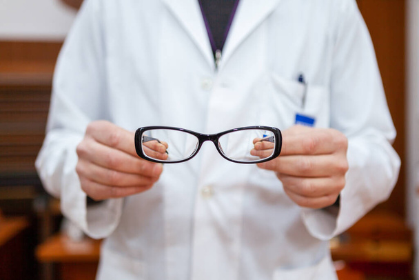 Een opticien in een witte vacht toont een paar corrigerende brillen om bijziendheid te corrigeren. De bril heeft een zwart frame en de handen van de opticien en de achtergrond zijn wazig. - Foto, afbeelding