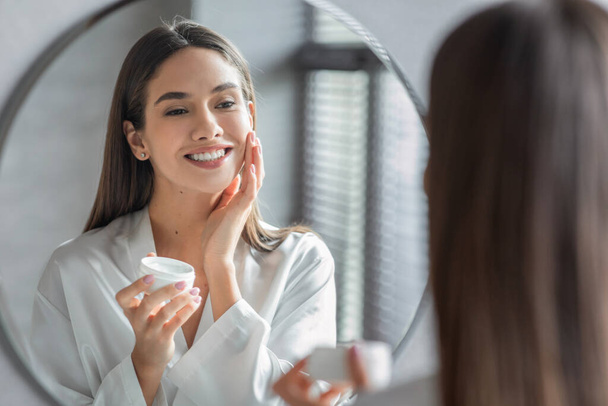Cosmetics Concept. Young Woman Looking At Mirror And Applying Face Cream - Foto, afbeelding