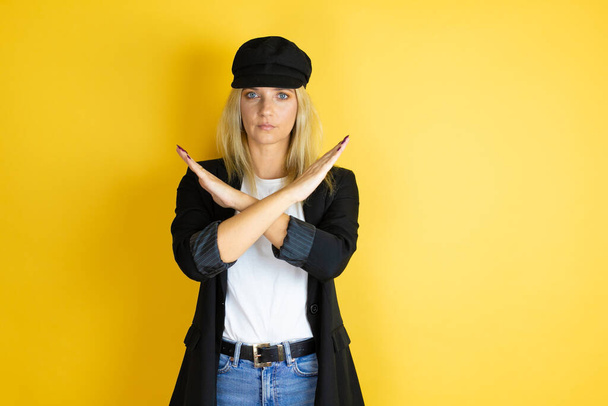 Beautiful woman wearing casual white t-shirt and a cap over isolated yellow background Rejection expression crossing arms doing negative sign, angry face - Photo, Image