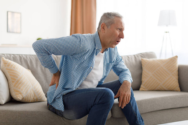 Dojrzały mężczyzna z bólem pleców siedzi na kanapie w domu - Zdjęcie, obraz
