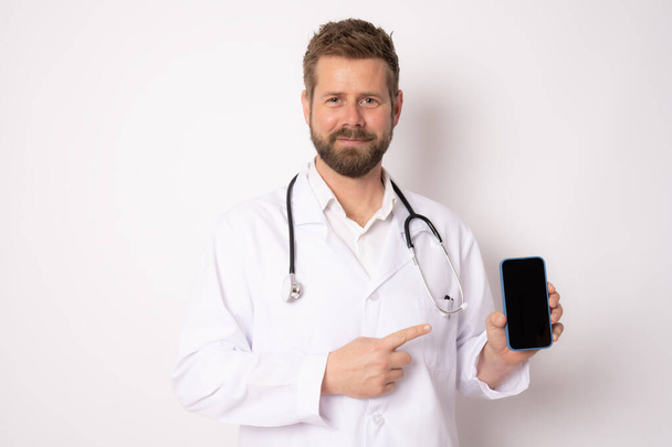Joven médico masculino sonriente que muestra la pantalla del teléfono móvil aislado sobre fondo blanco. Concepto médico. - Foto, imagen