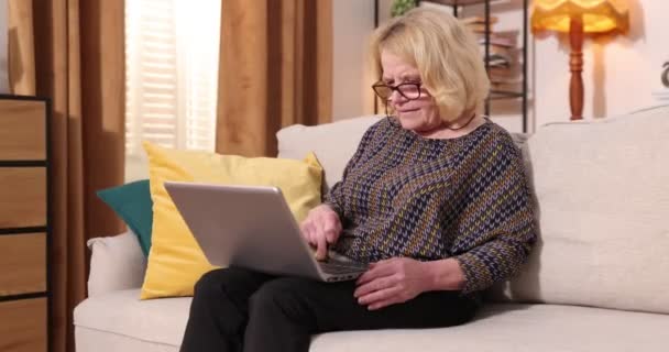 ポータブルコンピュータを使用して自宅でソファに座って灰色の髪のシニア女性の完全なビュー - 映像、動画