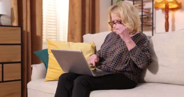 Plný pohled na šedovlasou starší ženu sedící doma na pohovce, používající přenosný počítač - Záběry, video