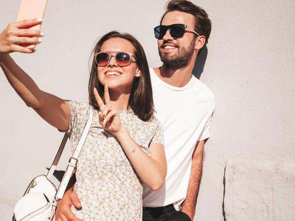 Smiling beautiful woman and her handsome boyfriend. Woman in casual summer clothes. Happy cheerful family. Female having fun. Couple posing in the street near wall.Hugging each other. Taking selfie - Zdjęcie, obraz
