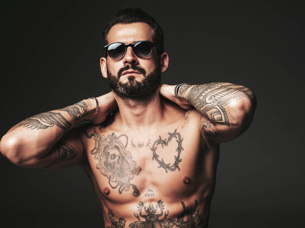 Portrait of handsome confident stylish hipster lambersexual model. Sexy modern man. Naked torso with tattoos.Fashion male posing in studio on dark background in sunglasses - Foto, imagen
