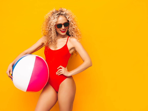 Young beautiful smiling woman posing near yellow wall in studio.Sexy model in red swimwear bathing suit.Positive female with curls hairstyle. Holding penny inflatable ball.Happy and cheerful - Фото, изображение