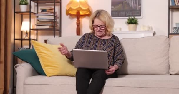 Pełny widok siwowłosej starszej kobiety siedzącej na kanapie w domu, korzystającej z komputera przenośnego - Materiał filmowy, wideo