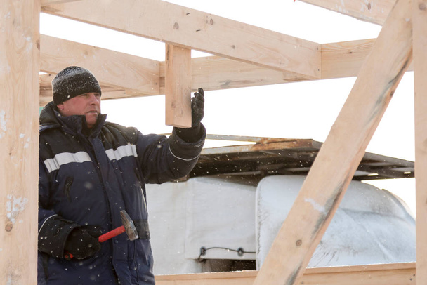 Budowniczy w niebieskiej kurtce z kapturem z deską w ręku na montażu drewnianej prowadnicy - Zdjęcie, obraz
