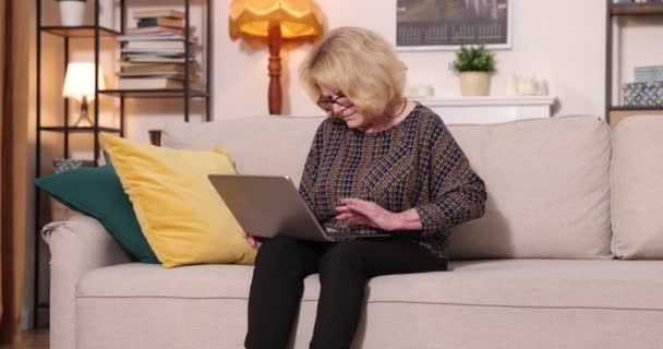 Vue complète de la femme âgée aux cheveux gris assise sur un canapé à la maison, à l'aide d'un ordinateur portable - Séquence, vidéo
