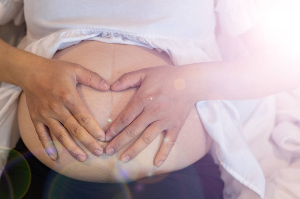Die werdende Mutter legt ihre Hände auf ihren Bauch, um mit ihrem ungeborenen Kind zu kommunizieren und auf den Tag zu warten, an dem ihr Baby geboren wird. Kommunikationskonzept von der Schwangeren bis zum Ungeborenen. - Foto, Bild