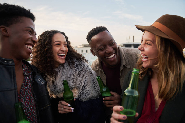 Laughing group of multi-cultural young adults socializing together on a rooftop terrace in the city. Drinks in hand, smiles with friends. - Фото, изображение