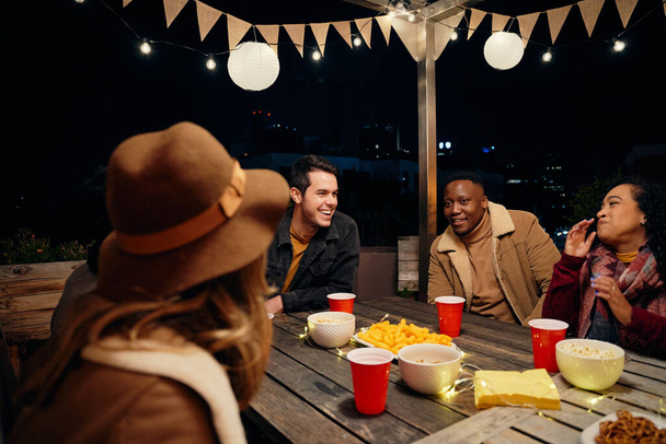 Multi-cultural group of young adult friends laughing while around a table at a trendy rooftop party. Eating and drinking, having a good time - Foto, imagen