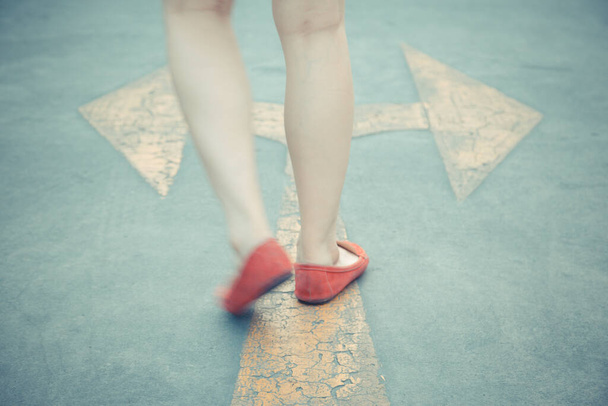 Vintage tono de niña usan zapatos rojos caminando hacia con señalización de flecha de tráfico amarillo en un fondo de carretera de asfalto - Foto, imagen