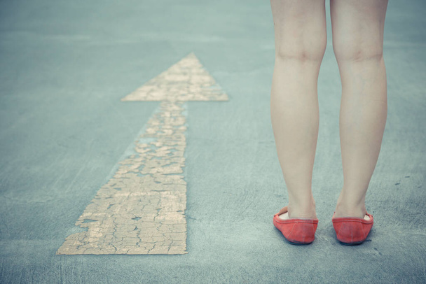 Vintage tono de niña usan zapatos rojos caminando hacia con señalización de flecha de tráfico amarillo en un fondo de carretera de asfalto - Foto, imagen