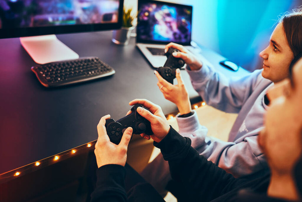 Friends playing video game at home. Gamers holding gamepads sitting at front of screen. Streamers girl and boy playing online in dark room lit by neon lights. Competition and having fun - Zdjęcie, obraz