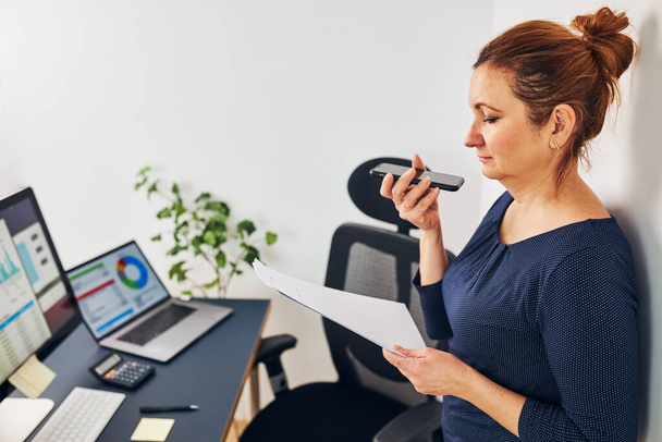 Woman entrepreneur having business  conversation on mobile phone. Businesswoman working in office holding documents and smartphone. Woman standing by desk with computers - Photo, Image