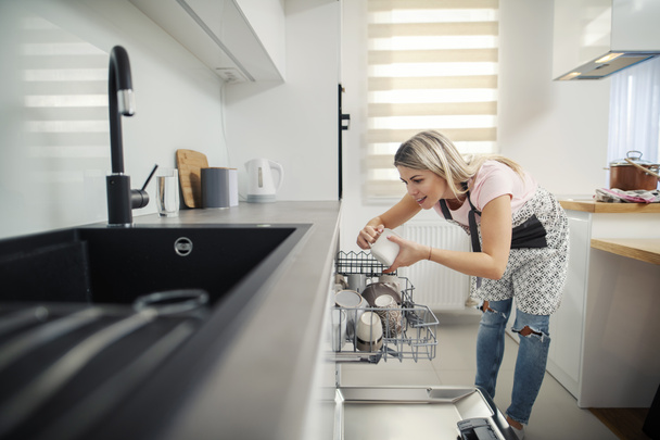 Czysta kobieta wkładająca naczynia do zmywarki w kuchni. - Zdjęcie, obraz