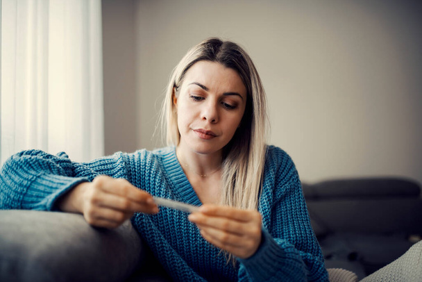 Uma mulher doente olhando para o termômetro enquanto se senta no sofá em sua casa. - Foto, Imagem
