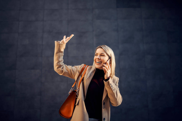 Una mujer de negocios feliz hablando por teléfono y saludando a un amigo mientras camina al aire libre. - Foto, imagen