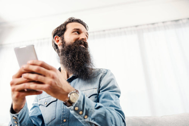 Vista de ángulo bajo de un hombre barbudo sosteniendo el teléfono y mirando hacia otro lado. - Foto, Imagen