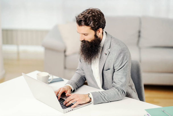 Um empreendedor barbudo focado sentado em casa e digitando no laptop. - Foto, Imagem