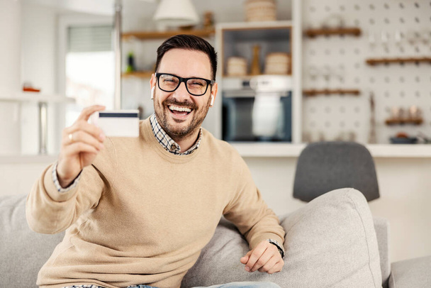 Un hombre feliz sentado en su acogedora casa y mostrando la tarjeta de crédito, el hombre está listo para las compras en línea. - Foto, Imagen