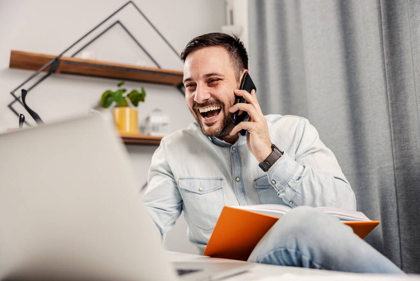 Un empresario riendo, hablando por teléfono y mirando el portátil y trabajando en su oficina en casa. - Foto, Imagen