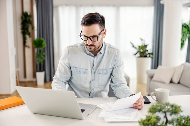 Um homem em sua casa acolhedora olhando para o laptop, trabalhando em papelada e resolve as finanças domésticas. - Foto, Imagem