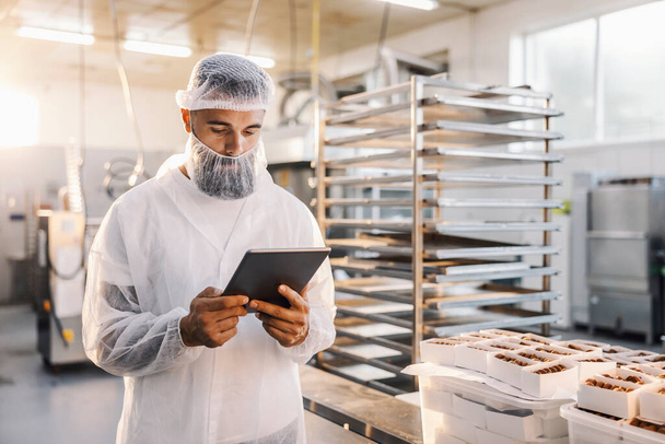 Εργάτης σε εργοστάσιο τροφίμων που χρησιμοποιεί tablet και ελέγχει τα cookies. - Φωτογραφία, εικόνα