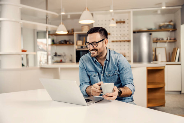 Ein glücklicher Mann, der Kaffee trinkt und in seinem gemütlichen Zuhause auf den Laptop schaut. - Foto, Bild