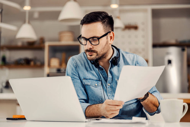 Un entrepreneur concentré regardant l'ordinateur portable et travaillant sur la paperasserie à sa maison confortable. - Photo, image