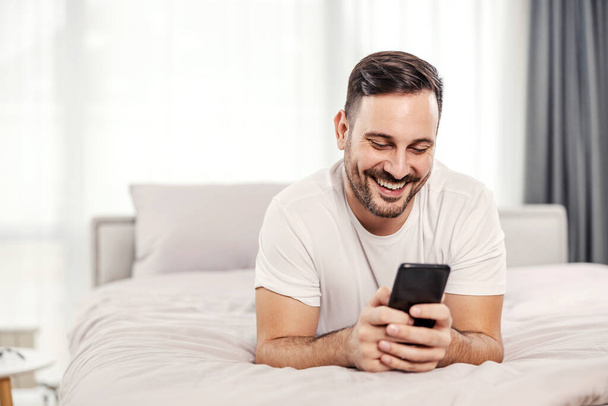Ένας χαρούμενος πρωινός άντρας ξαπλωμένος στο κρεβάτι και φλερτάροντας στο τηλέφωνο στο άνετο διαμέρισμά του. - Φωτογραφία, εικόνα