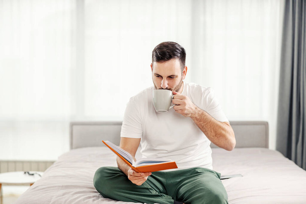 Um homem da manhã bebendo seu café e lendo um livro na cama em seu aconchegante apartamento. - Foto, Imagem