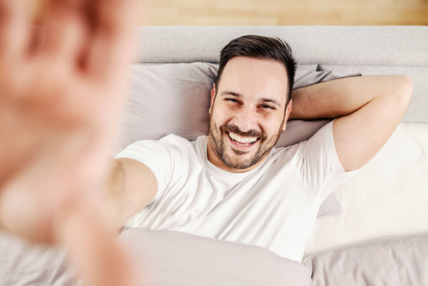 Ένας ευτυχισμένος άντρας ξαπλωμένος στο κρεβάτι και βγάζει σέλφι.. - Φωτογραφία, εικόνα