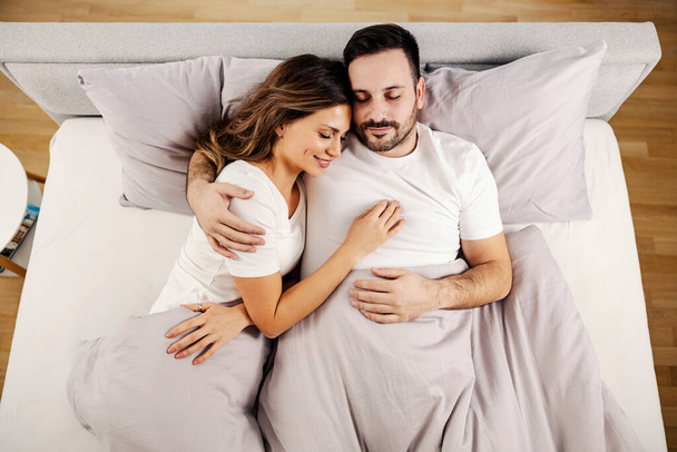 Κάτοψη ενός ευτυχισμένου πρωινού ζευγαριού ξαπλωμένο στο κρεβάτι τους, αγκαλιασμένο στο άνετο σπίτι τους. - Φωτογραφία, εικόνα