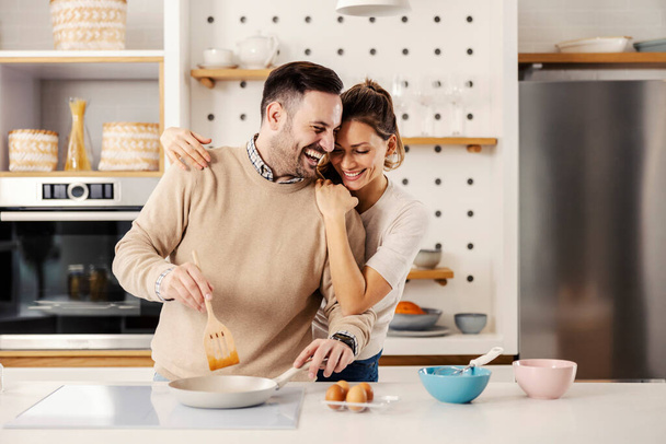Un homme dans la cuisine prépare un repas pendant que sa femme le serre dans ses bras. - Photo, image