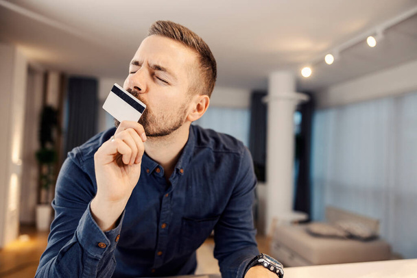 Mężczyzna całujący swoją nową kartę kredytową i wydający pieniądze online w domu. - Zdjęcie, obraz