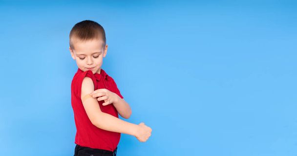 Zadowolony chłopiec na ramieniu z plastrem na ramieniu po szczepieniu na niebieskim tle, Koncepcja szczepienia przeciwko koronawirusowi lub COVID-19. - Zdjęcie, obraz