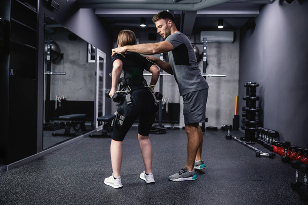 Un entrenador personal masculino joven ayuda a una mujer con un traje muscular eléctrico para estimular los músculos. Fitness en traje EMS en concepto de gimnasio moderno, entrenamiento y cuidados revolucionarios, estimulación eléctrica - Foto, Imagen