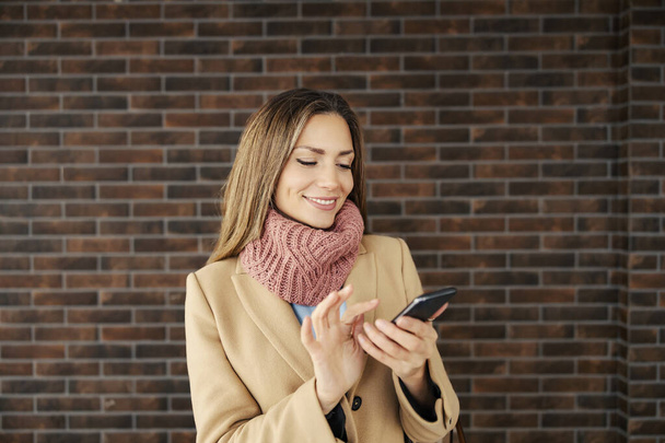 Egy nő áll az utcán és sms-ezik az okostelefonon. Egy fiatal boldog nő meleg ruhában áll az utcán a téglafal előtt, és SMS-t ír egy barátjának.. - Fotó, kép