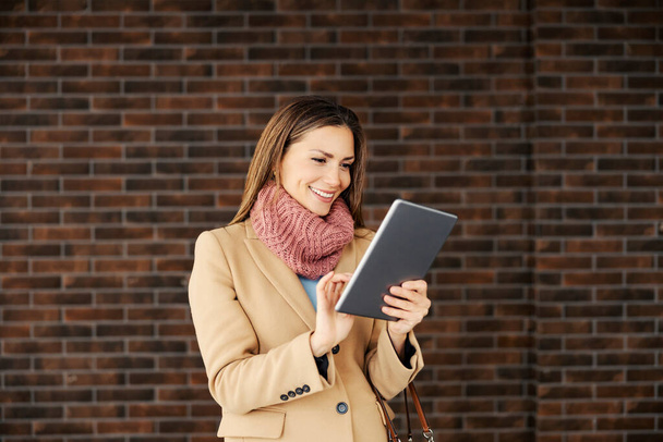 Geschäftsfrau mit Tablet im Freien.Eine junge fröhliche modische Geschäftsfrau in warmen Kleidern steht auf der Straße vor der Ziegelmauer und checkt mit einem Tablet E-Mails. - Foto, Bild
