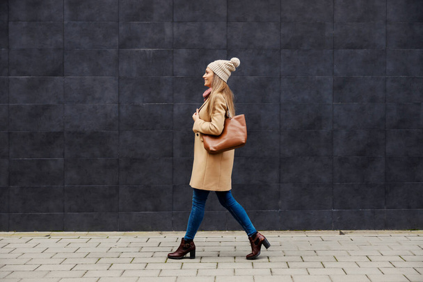 Modieus, stijlvol en fantastisch in de winter. Volledige lengte van een jonge gelukkige vrouw in jas, sjaal, en muts lopen op straat in koud weer. - Foto, afbeelding